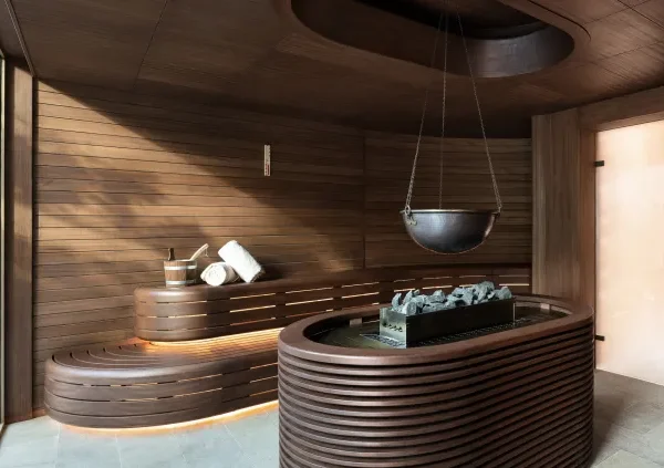 Modern wooden sauna with curved benches, towels, bucket, and central heater holding sauna stones.