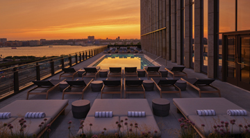 Sunset rooftop pool deck with lounge chairs, glass railings, and a pool reflecting the orange sky.
