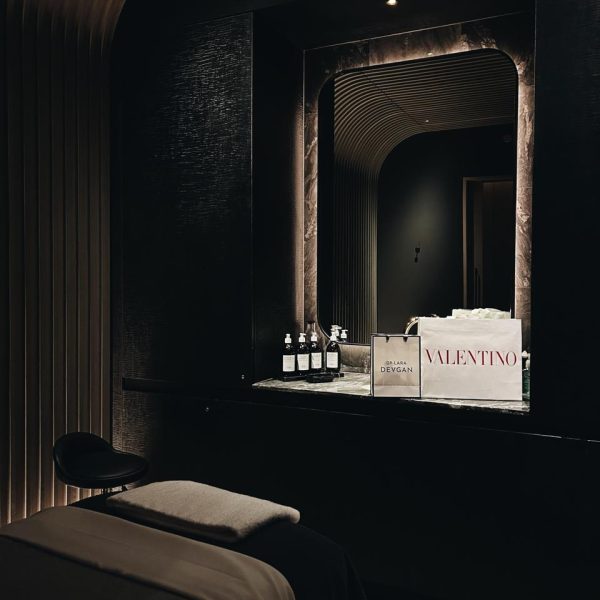 Dim luxurious spa with dark walls, large mirror, skincare bottles, shopping bags, and massage table.