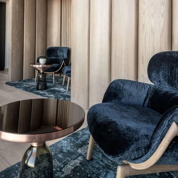 Seating area with dark blue velvet chairs, patterned rugs, light wood walls, and a metallic table.