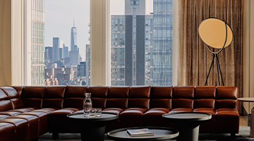 Lounge with brown leather sectional, round tables, big windows, skyline view, and tall floor lamp.
