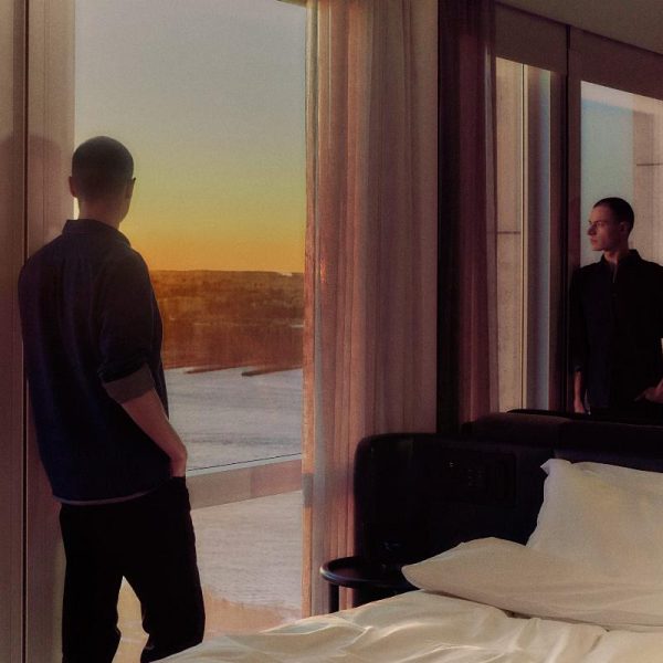 Person standing by tall windows at sunset with their reflection beside an unmade bed.