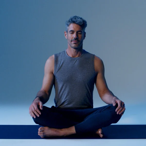 Man sitting cross‑legged on yoga mat, eyes closed, meditating in blue lighting.