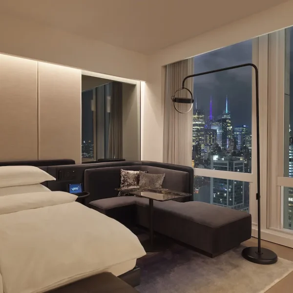 Modern hotel room with white bed, corner sofa, and tall windows revealing illuminated nighttime skyline.