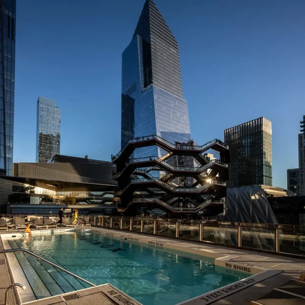 Rooftop pool with warm sunlight, tall glass towers, and a prominent honeycomb-style structure nearby.