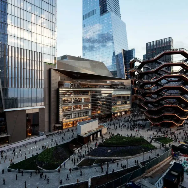Busy Hudson Yards plaza with the Vessel on the right and crowds moving among glass towers.