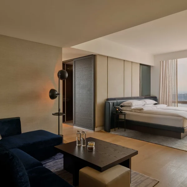 Modern open hotel suite with sofa, coffee table, white-bedded room, and large windows beyond.