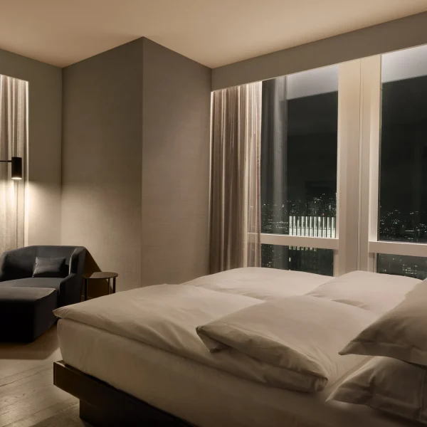 Modern hotel bedroom with white bedding, city-view windows, black armchair, side table, warm lighting.