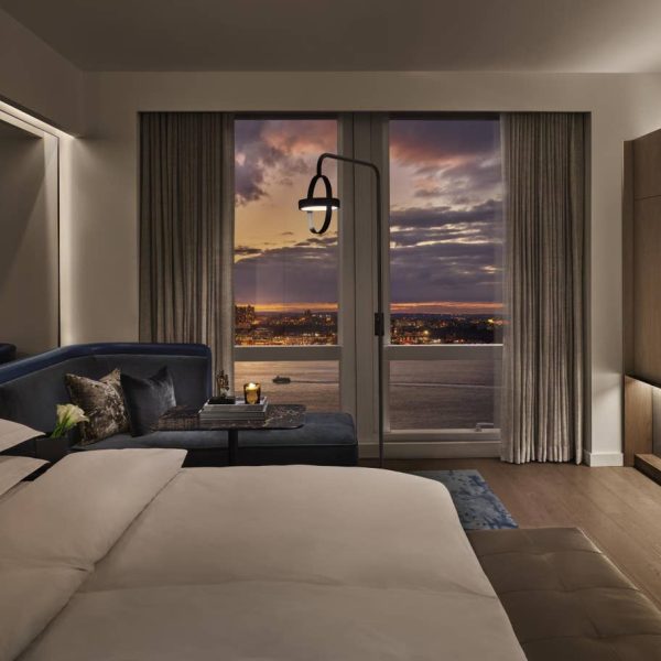 Hotel room with white bed and blue window sofa overlooking sunset, water, and city lights.