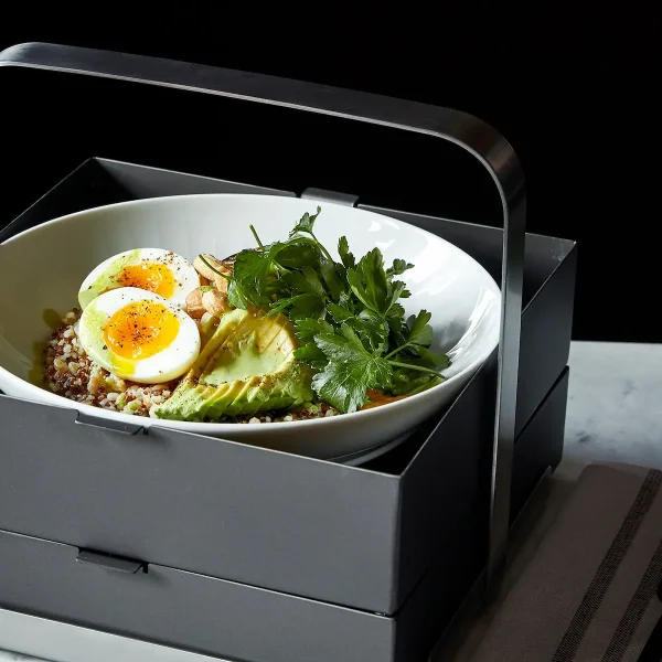 Breakfast bowl with soft‑boiled eggs, avocado, herbs, and grains in modern gray tray.