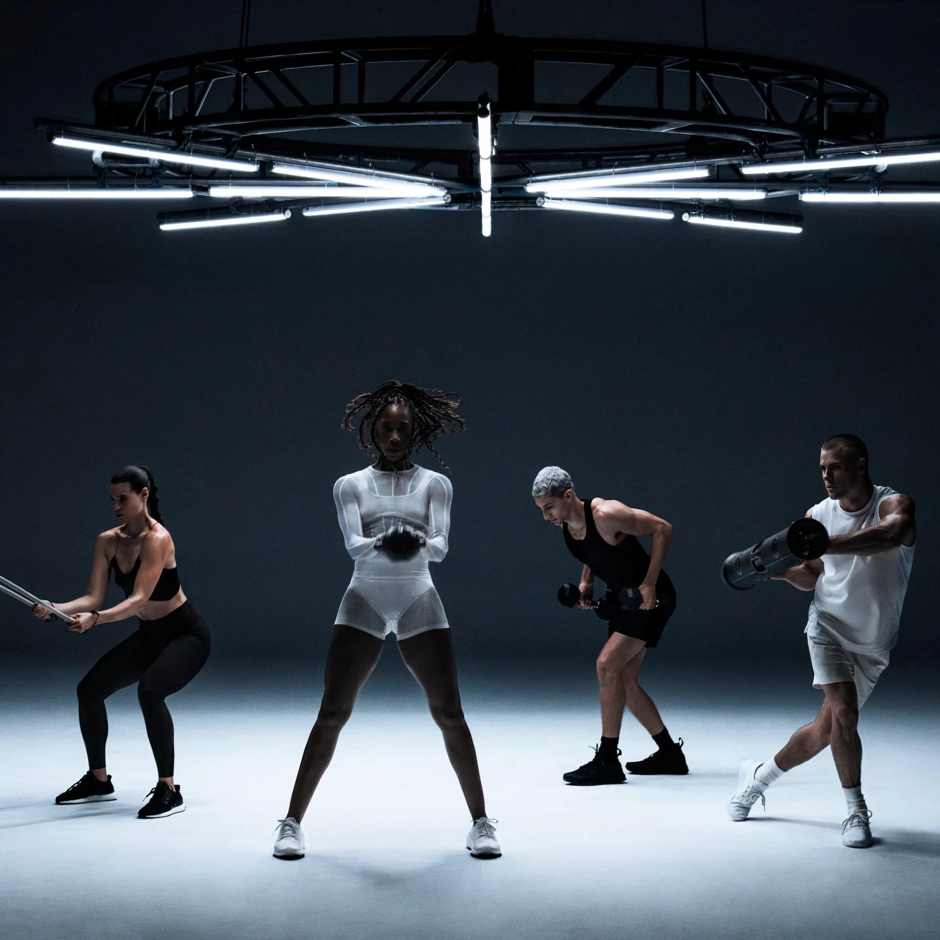 Four individuals performing strength exercises under radial overhead lights in dark, minimalist fitness space