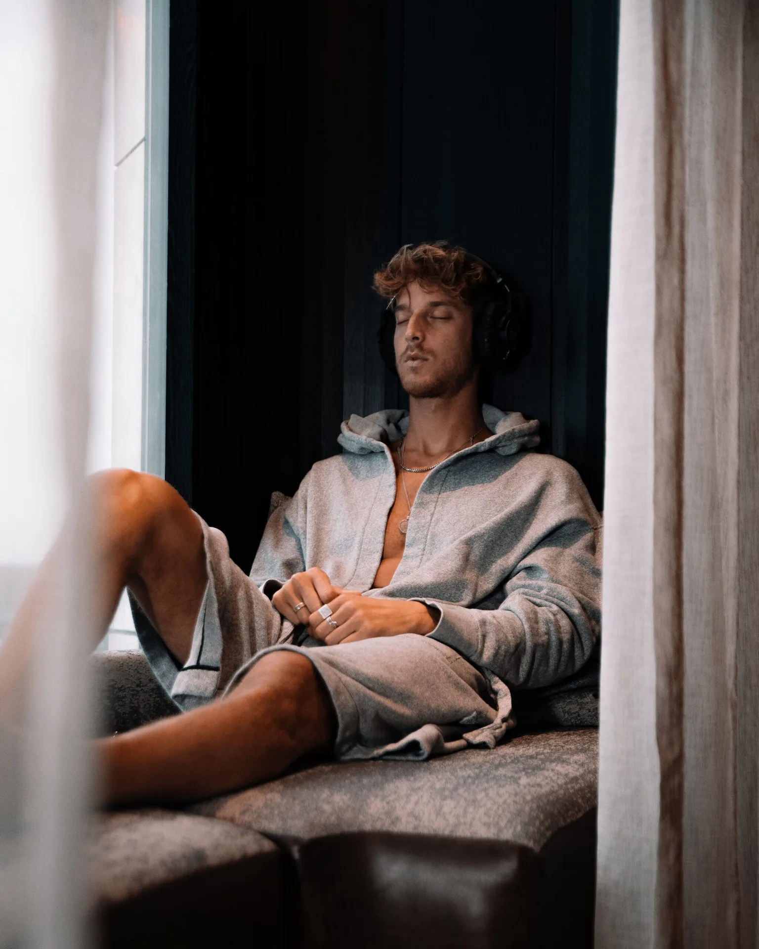 Person in gray hoodie and shorts sitting by window with headphones, eyes closed in relaxed wellness moment