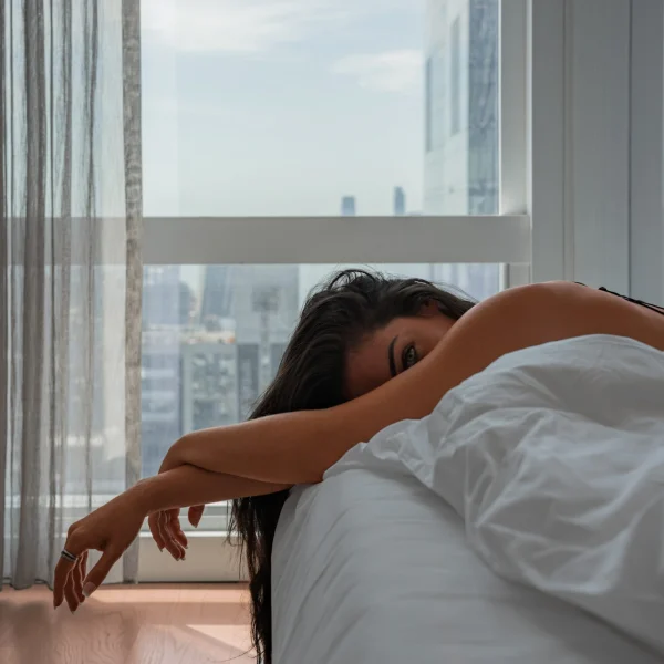 Person resting on bed with white blanket in sunlit high-rise room overlooking city buildings