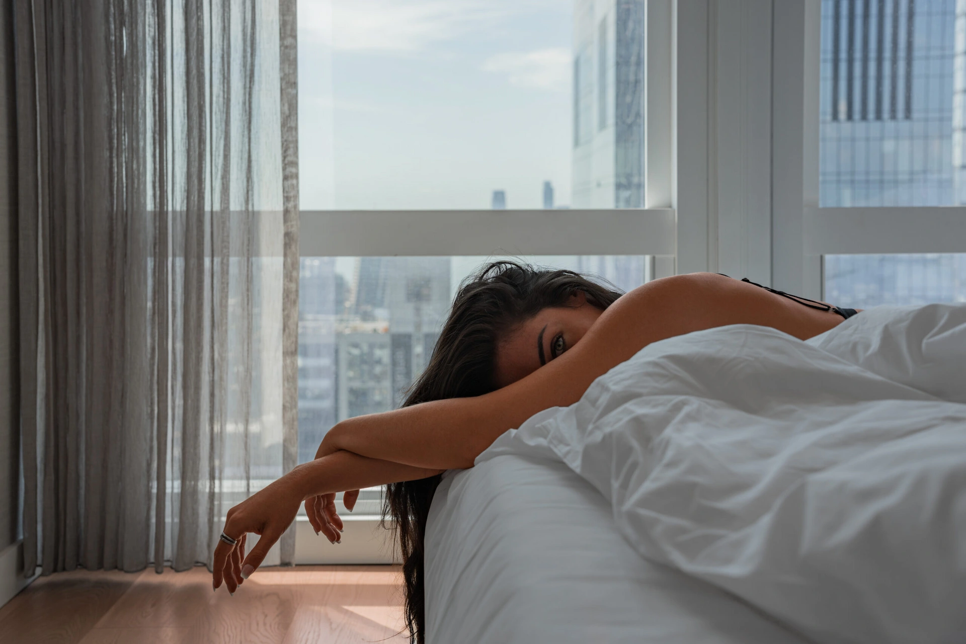 Person resting on bed with white blanket in sunlit high-rise room overlooking city buildings