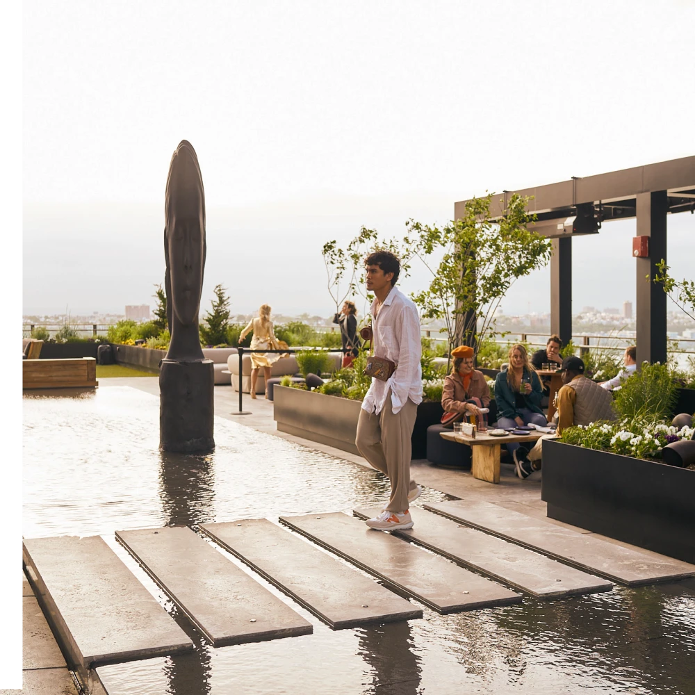 Man walking on rooftop stone path over shallow pool with sculpture, greenery, and city view at Equinox Hotels