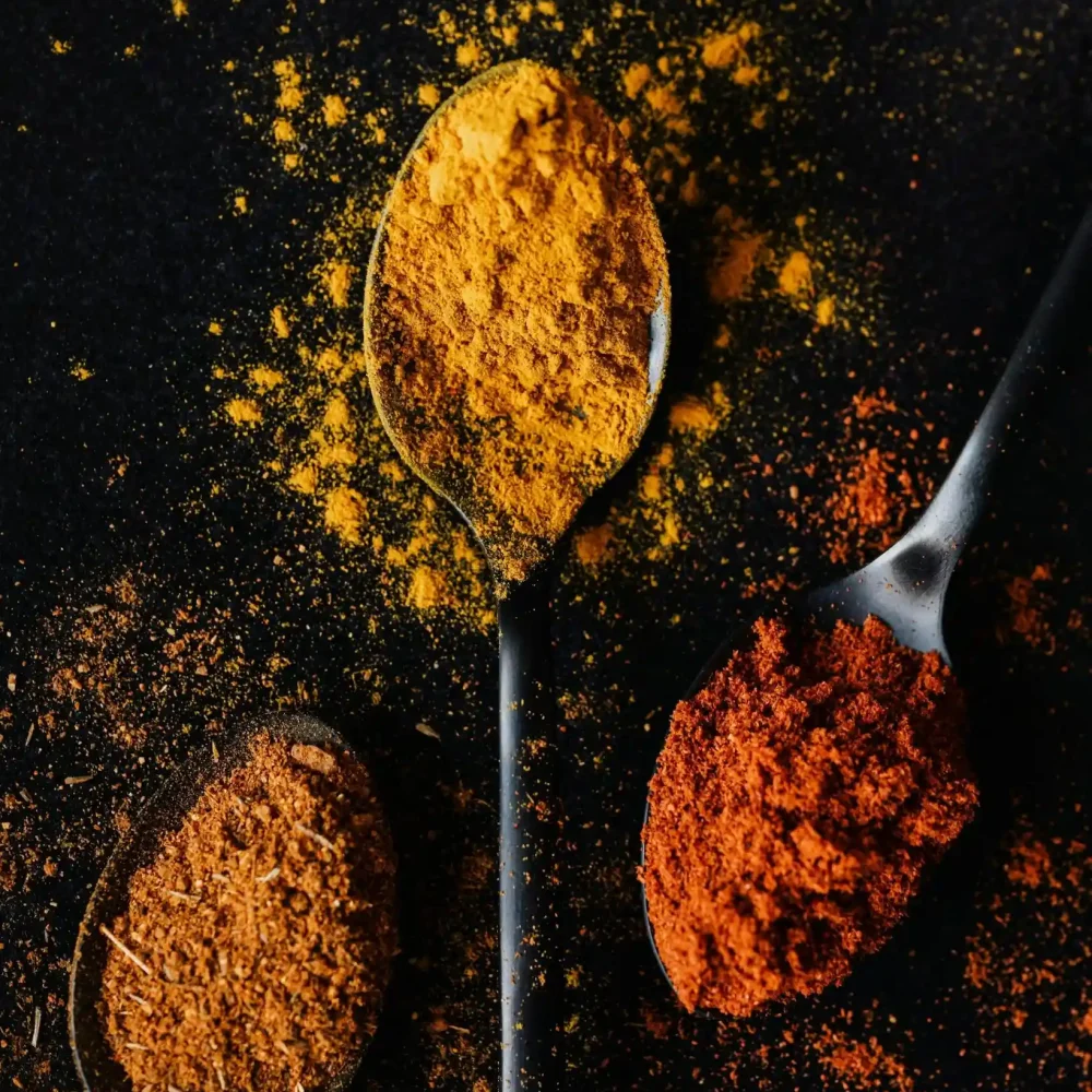 Three metal spoons on dark surface, each holding vibrant ground spices: turmeric cumin , and red paprika