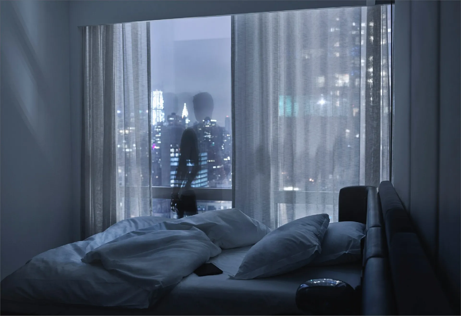 Silhouetted figure standing by window in dim bedroom with unmade bed and NYC skyline at night
