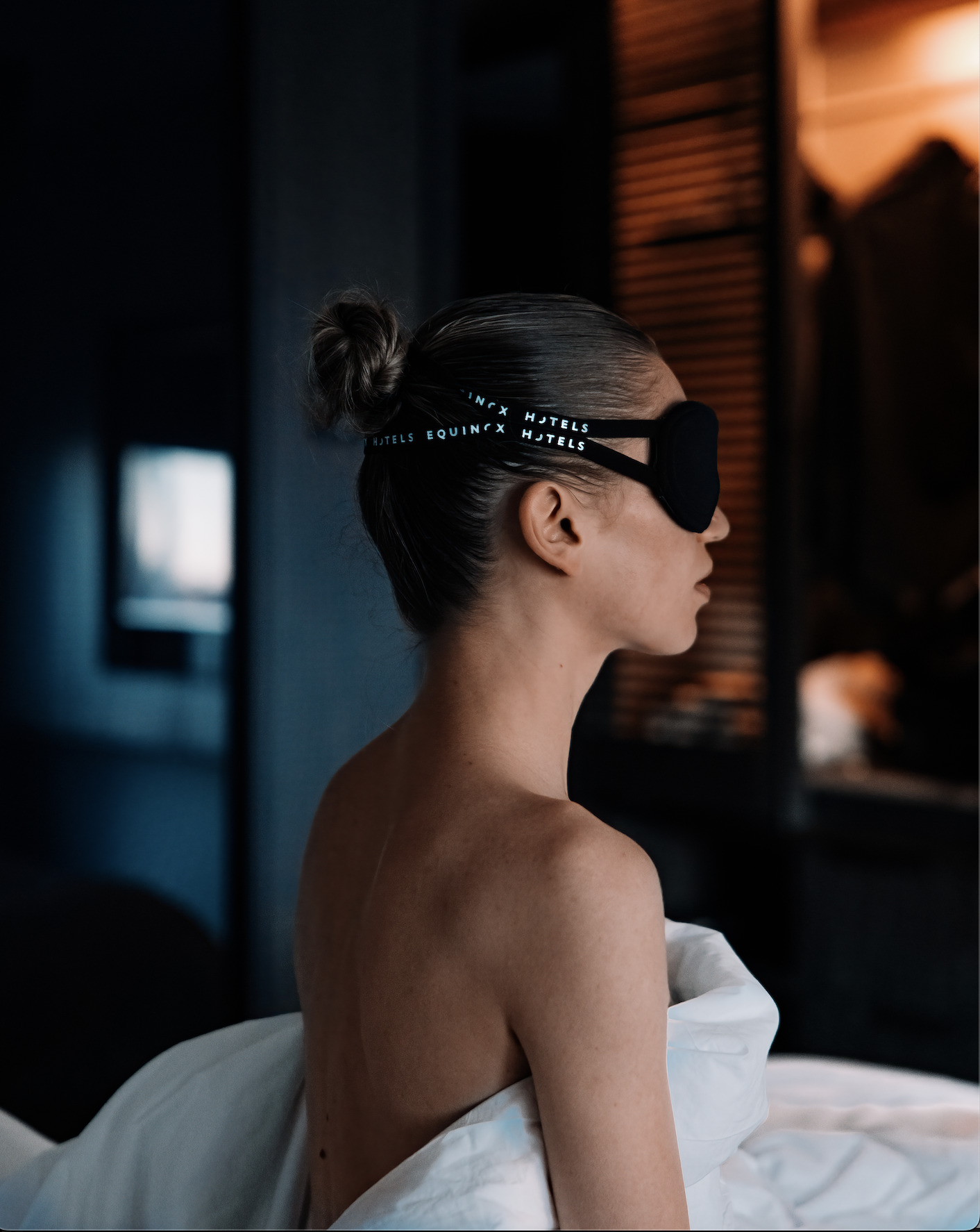 Woman sitting up in bed at Equinox Hotel, wearing a branded sleep mask and wrapped in white sheets, with only her shoulders and above visible as the rest of the room fades into a soft blur, highlighting a moment of restful stillness.