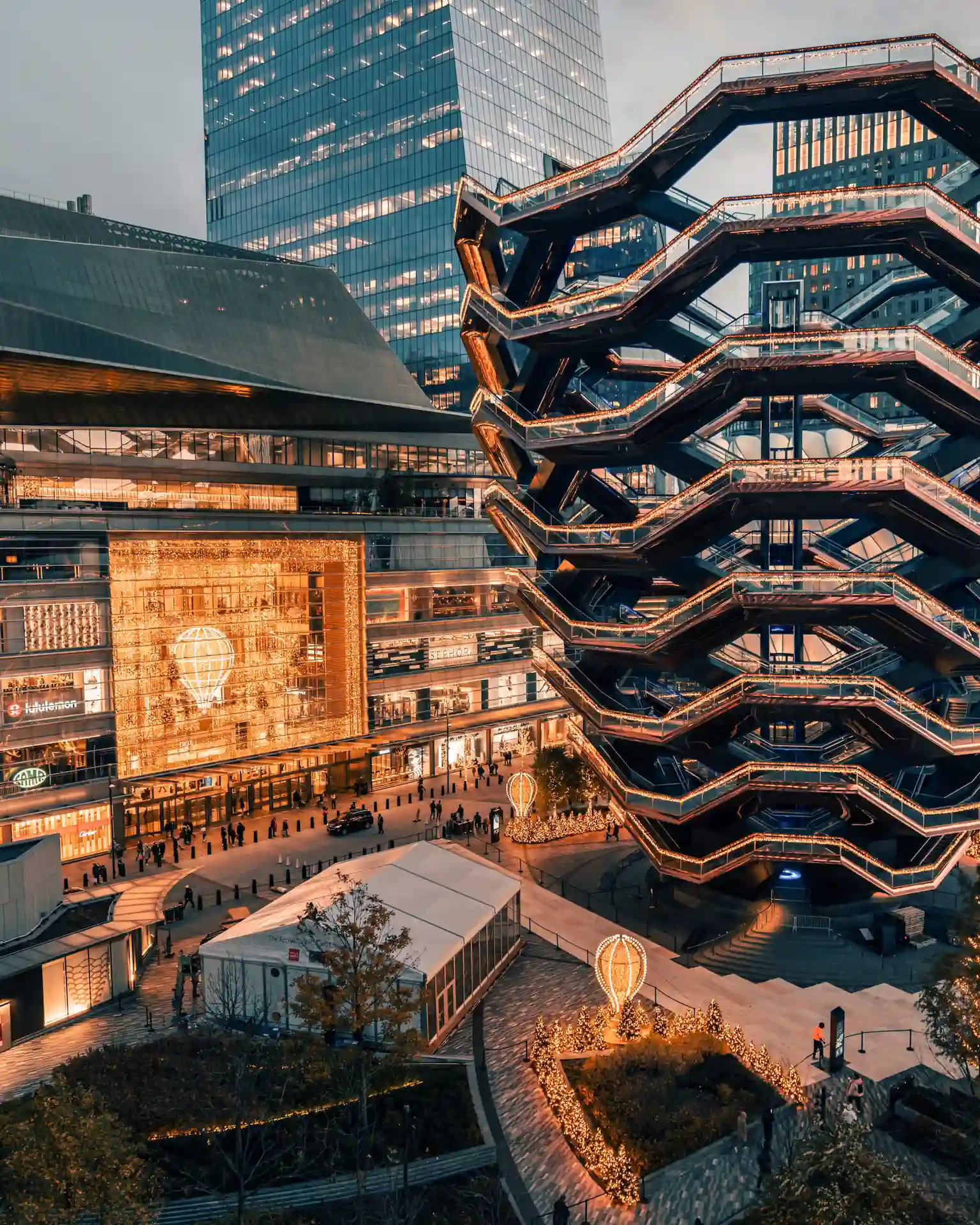 The Vessel at Hudson Yards lit with warm lights, surrounded by skyscrapers, people, and festive balloon displays.