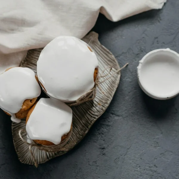 Three iced muffins on leaf-shaped plate with bowl of white frosting on dark textured surface