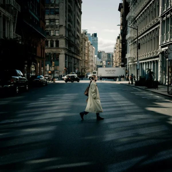 Person in beige coat crossing wide city street with dramatic shadows and historic buildings in background