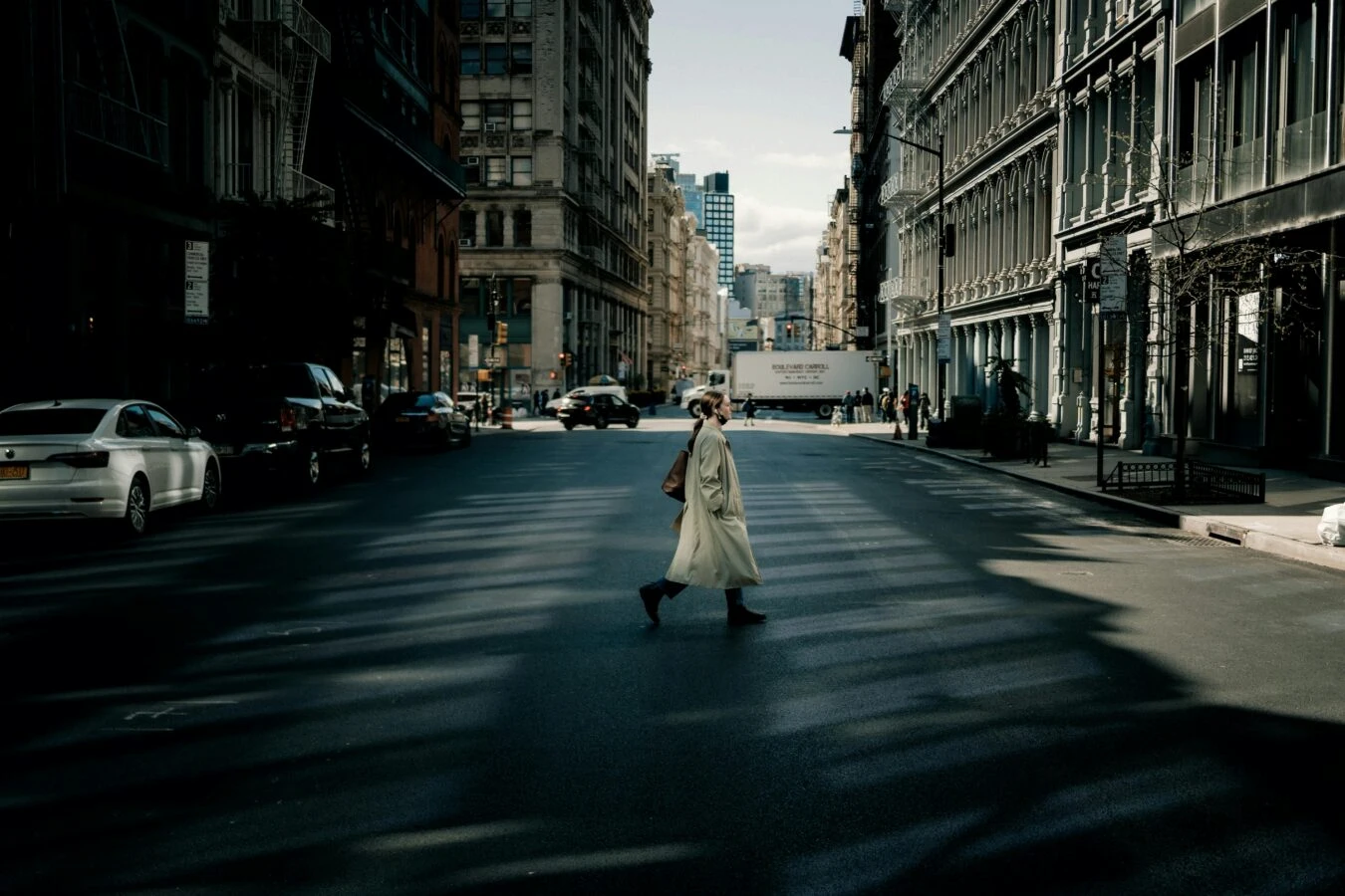 Person in beige coat crossing wide city street with dramatic shadows and historic buildings in background