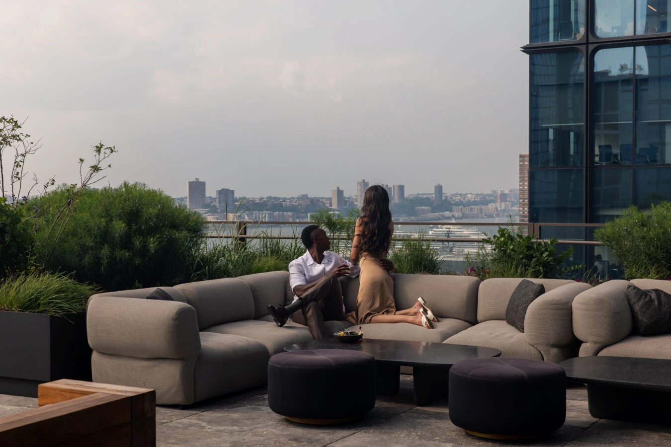 Two people on curved rooftop sofa with coffee table, greenery, and riverfront city view in background