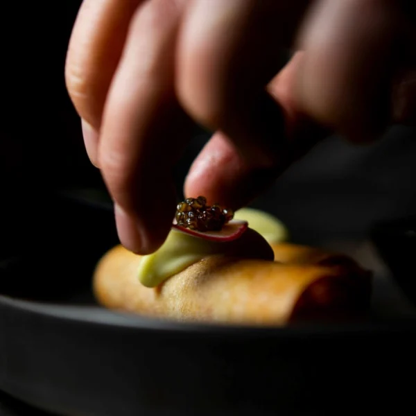 Close-up of hand placing caviar garnish on gourmet roll with radish and green vegetable on dark plate
