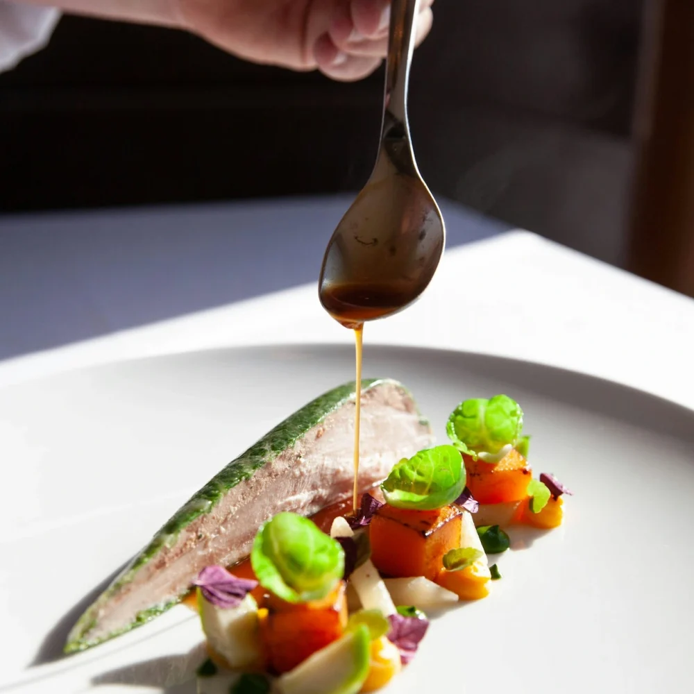 Equinox gourmet dish being plated with herb-crusted fish, roasted vegetables, greens, and brown sauce drizzle