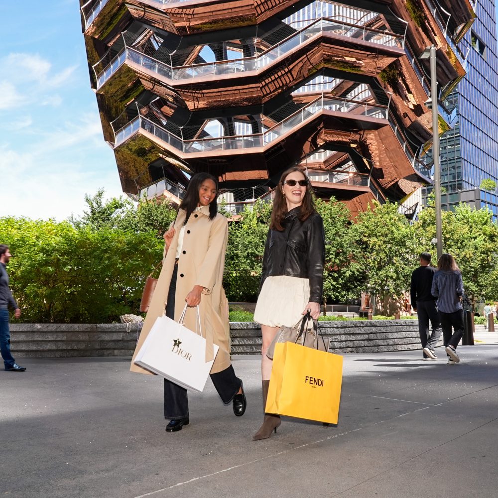 Two people with Dior and Fendi bags walking past the Vessel in Hudson Yards on a sunny day