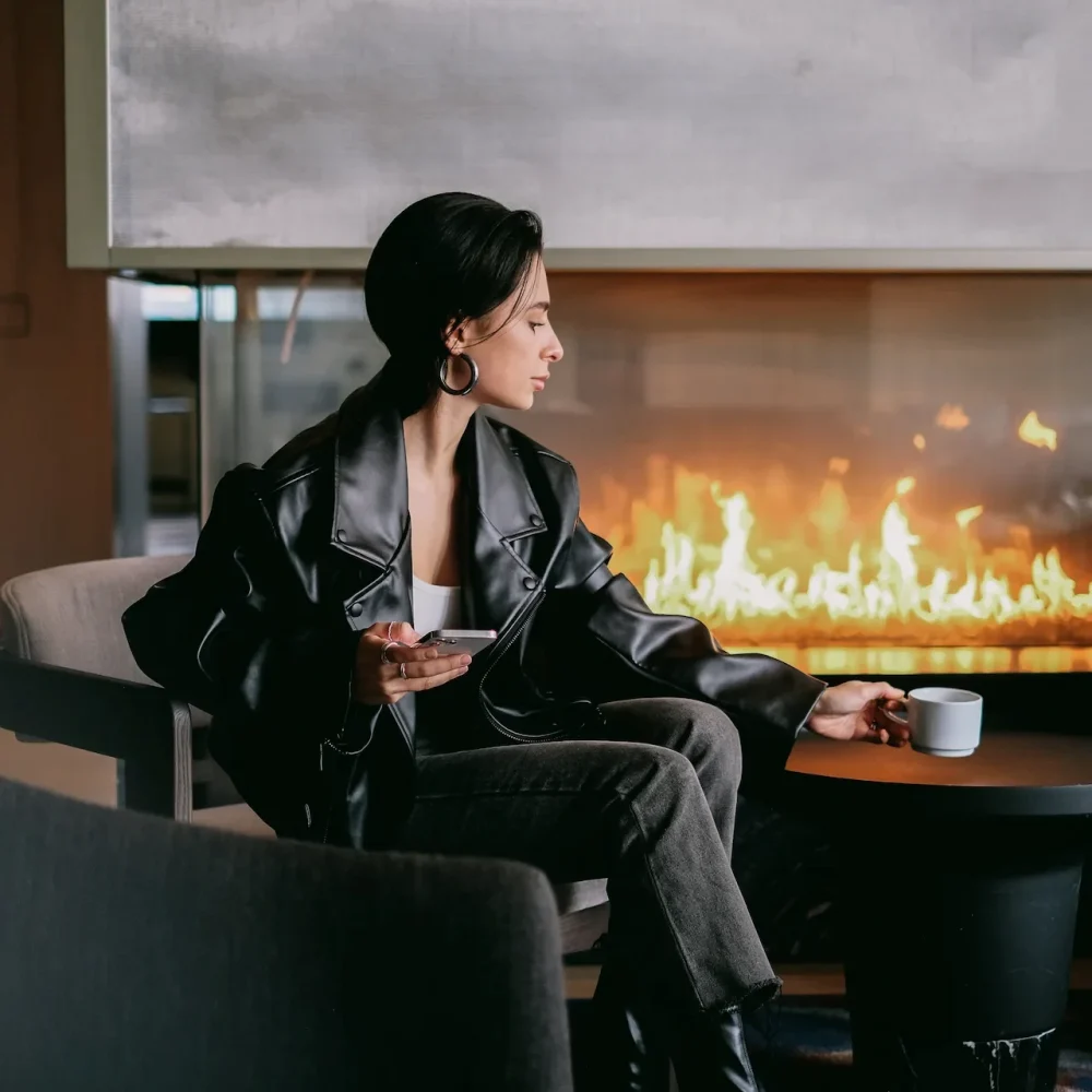 Person in black boots and leather jacket reaching for mug by fireplace in cozy modern lounge