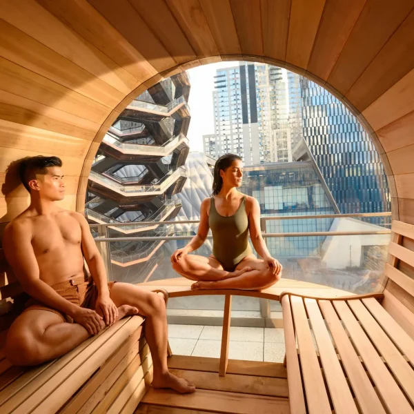 Two people relaxing inside circular wooden sauna with round window overlooking Vessel and NYC skyline