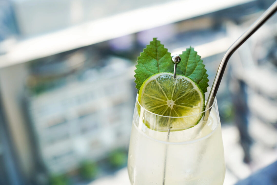 Close-up of cocktail with lime slice and green leaves in clear glass with metal straw on rooftop