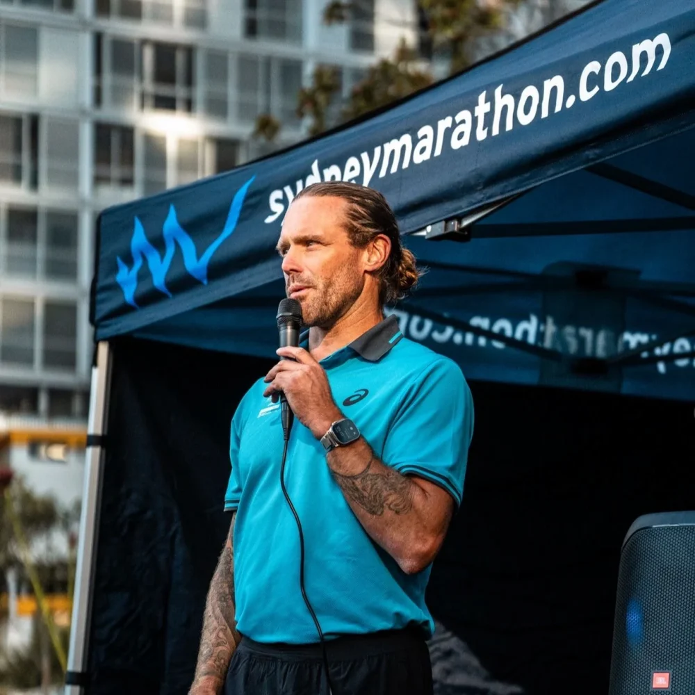 Man with tattoos speaking into mic at Sydney Marathon event under branded tent near modern building