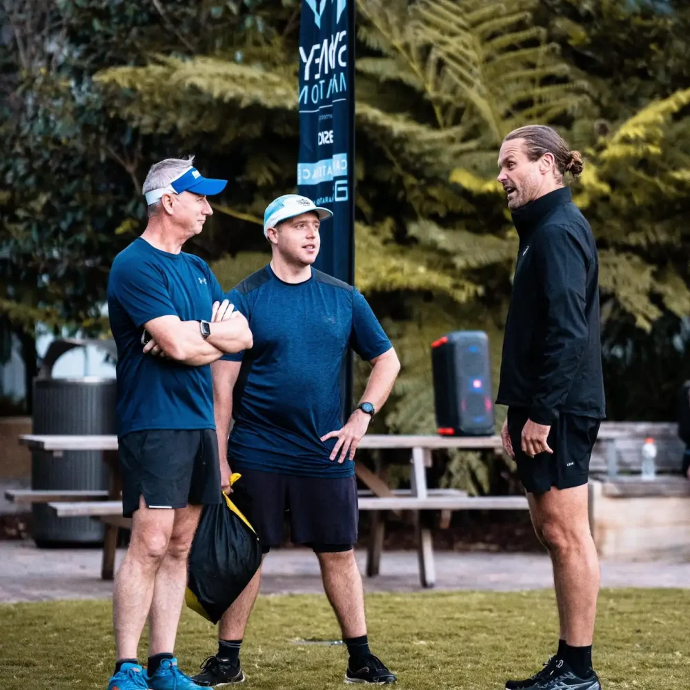 Three people in athletic gear talking on grassy area with trees, picnic table, and outdoor fitness setup