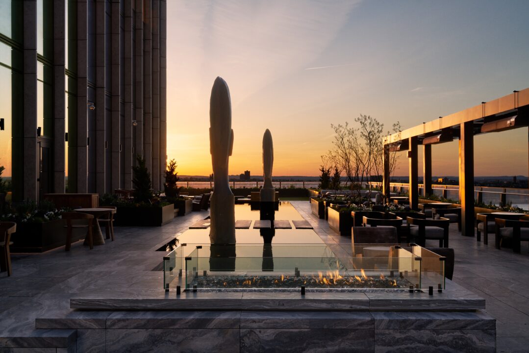 Rooftop terrace at sunset with sculptures, fire pit, seating and water views in elegant urban setting