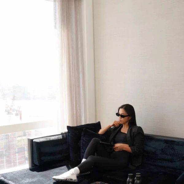 Person seated on blue velvet sofa with tablet and sunglasses in sunlit room overlooking urban construction