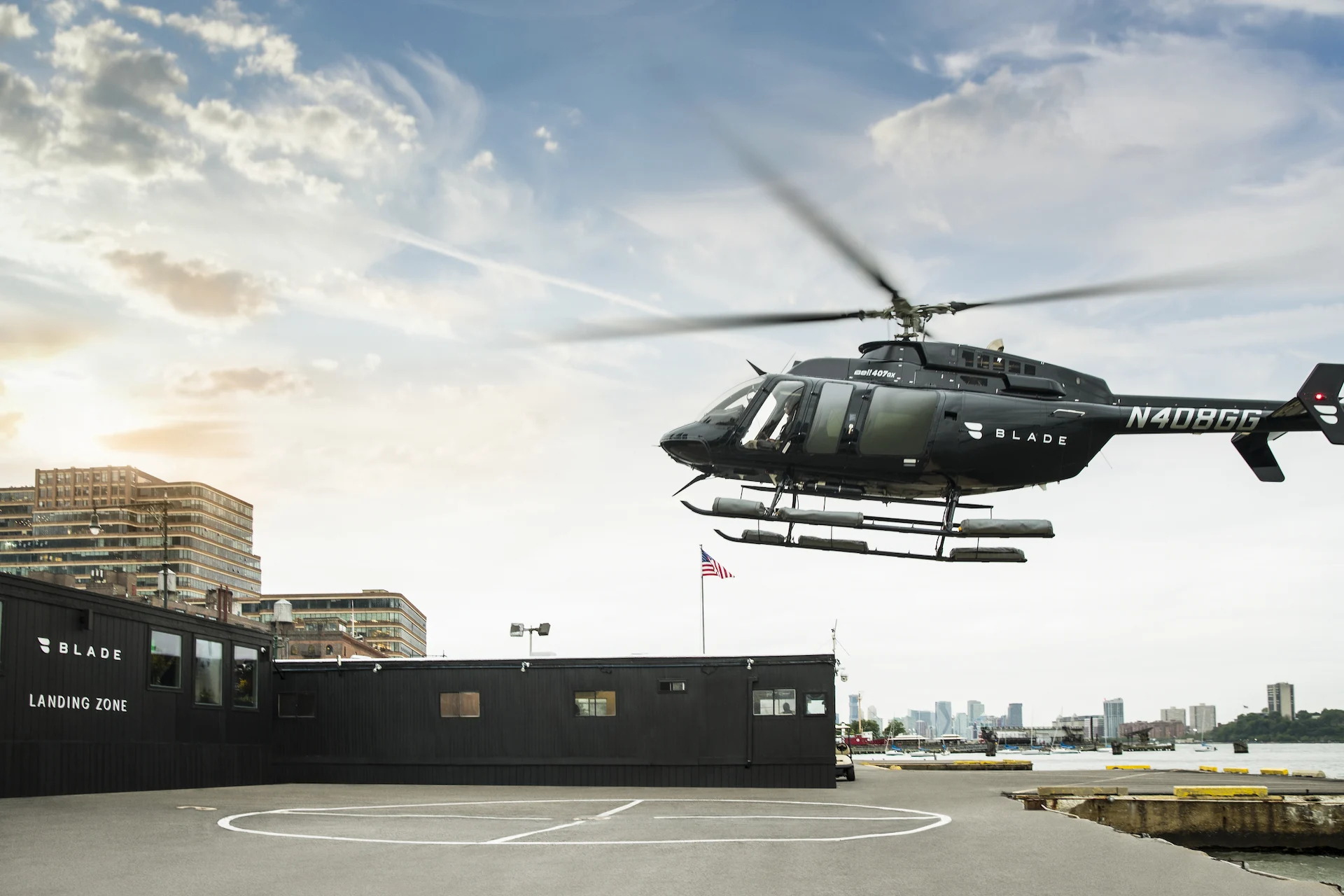 Black BLADE helicopter hovering over waterfront helipad near city skyline and landing zone building