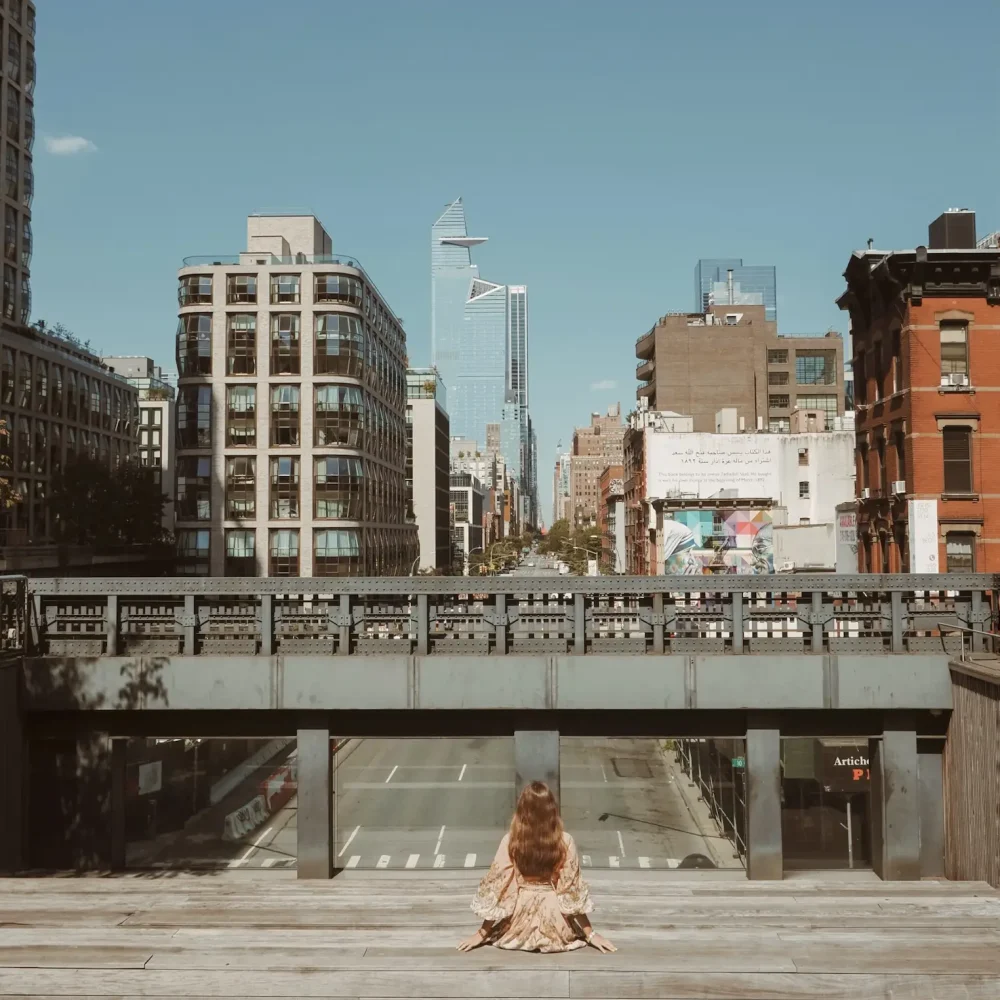 Person in patterned dress seated alone on wooden platform overlooking city skyline with mix of architecture
