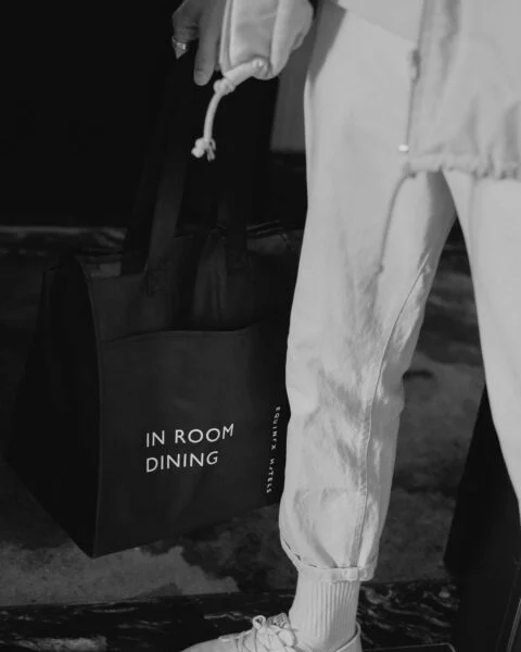 Person in white sneakers holding black “IN ROOM DINING” tote at Equinox, in elegant black-and-white hotel setting