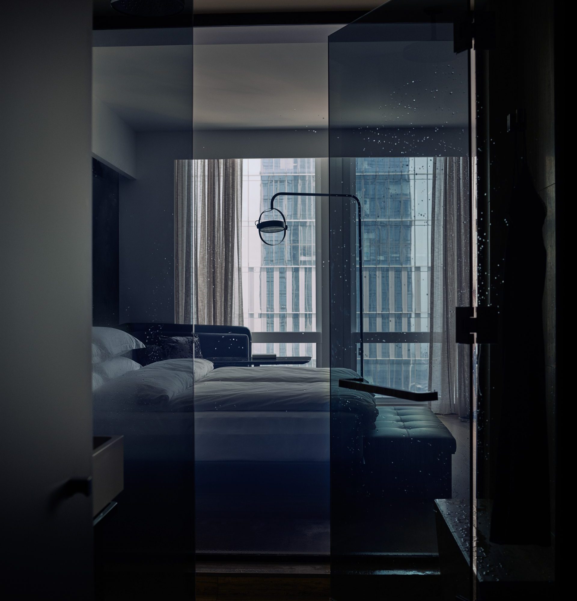 View of a sleek bedroom through wet glass, with white bed, bench, lamp, and city buildings outside window.