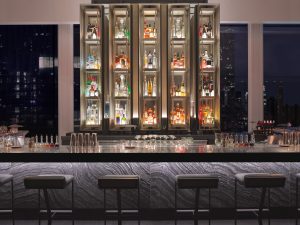 Upscale bar interior at night with sleek counter, high stools, and backlit liquor shelves against city skyline.