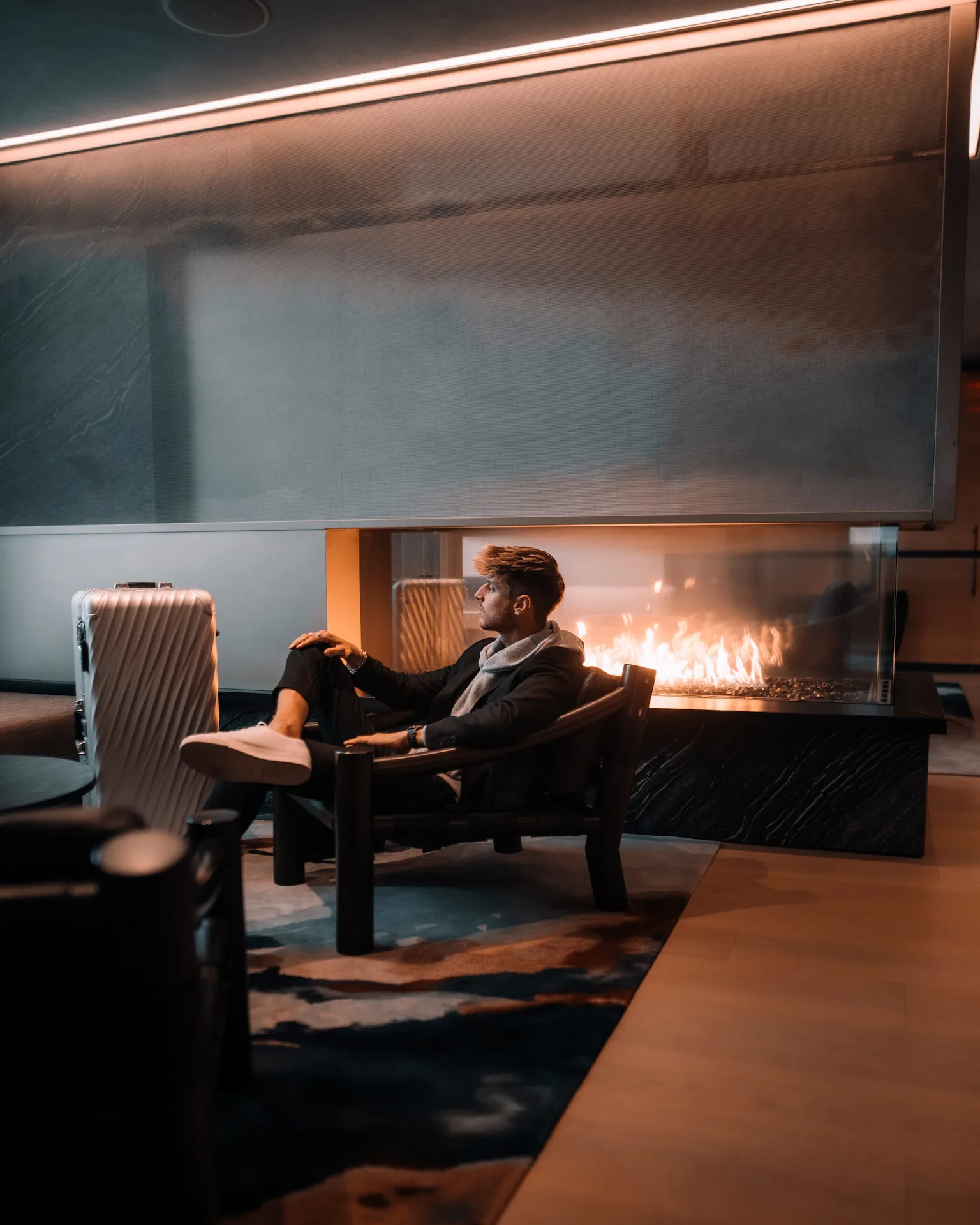Person in dark suit seated by sleek fireplace at Equinox hotel lounge with suitcase and warm lighting