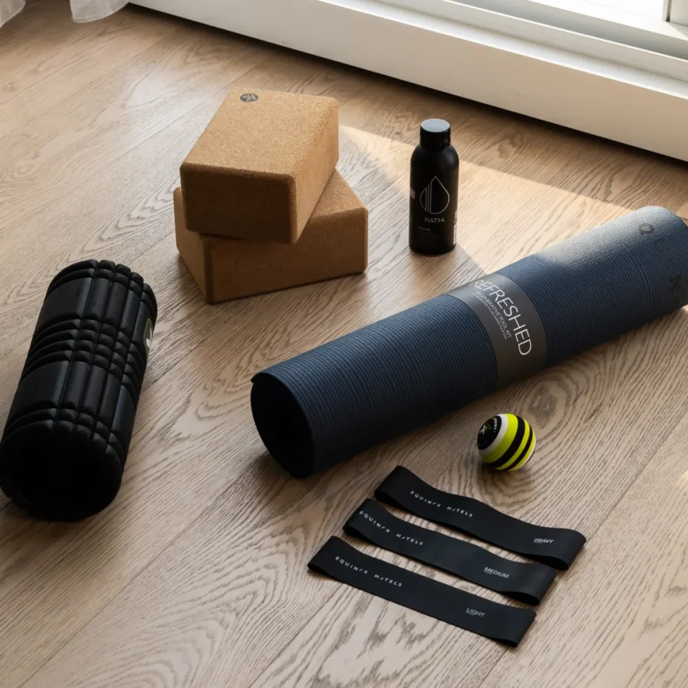 Fitness gear arranged on wooden floor near window, including yoga mat, cork blocks, foam roller, and resistance bands