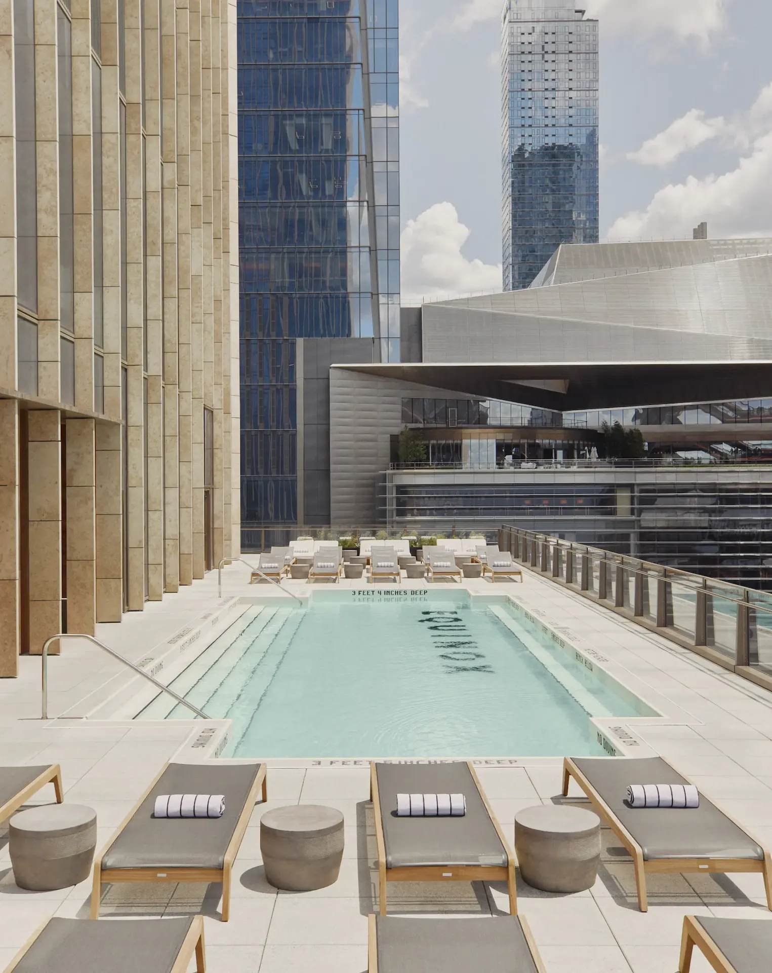 Rooftop pool with cushioned lounge chairs, clear blue water, and surrounding city skyscrapers