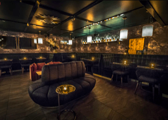Dimly lit lounge with curved sofa, pendant lights, textured walls, and a stocked bar in the background