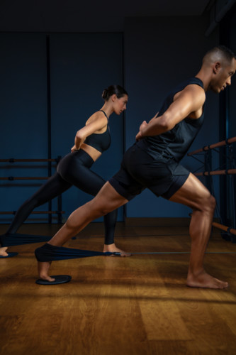 Two people in black athletic wear doing resistance band lunges in mirrored studio with wood floors