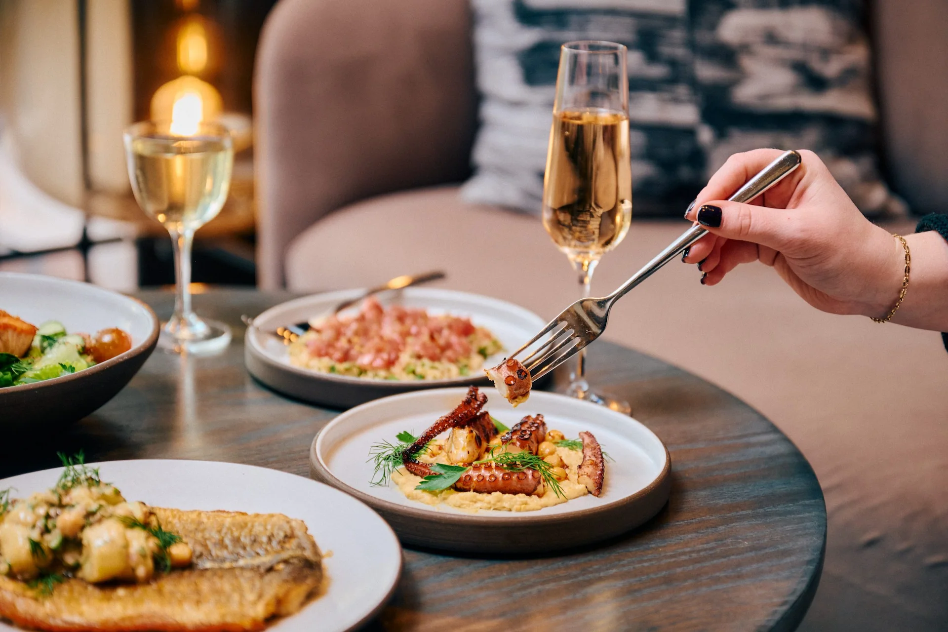 A diner uses a fork to eat grilled octopus at Electric Lemon, with glasses of wine and various small plates on the table.