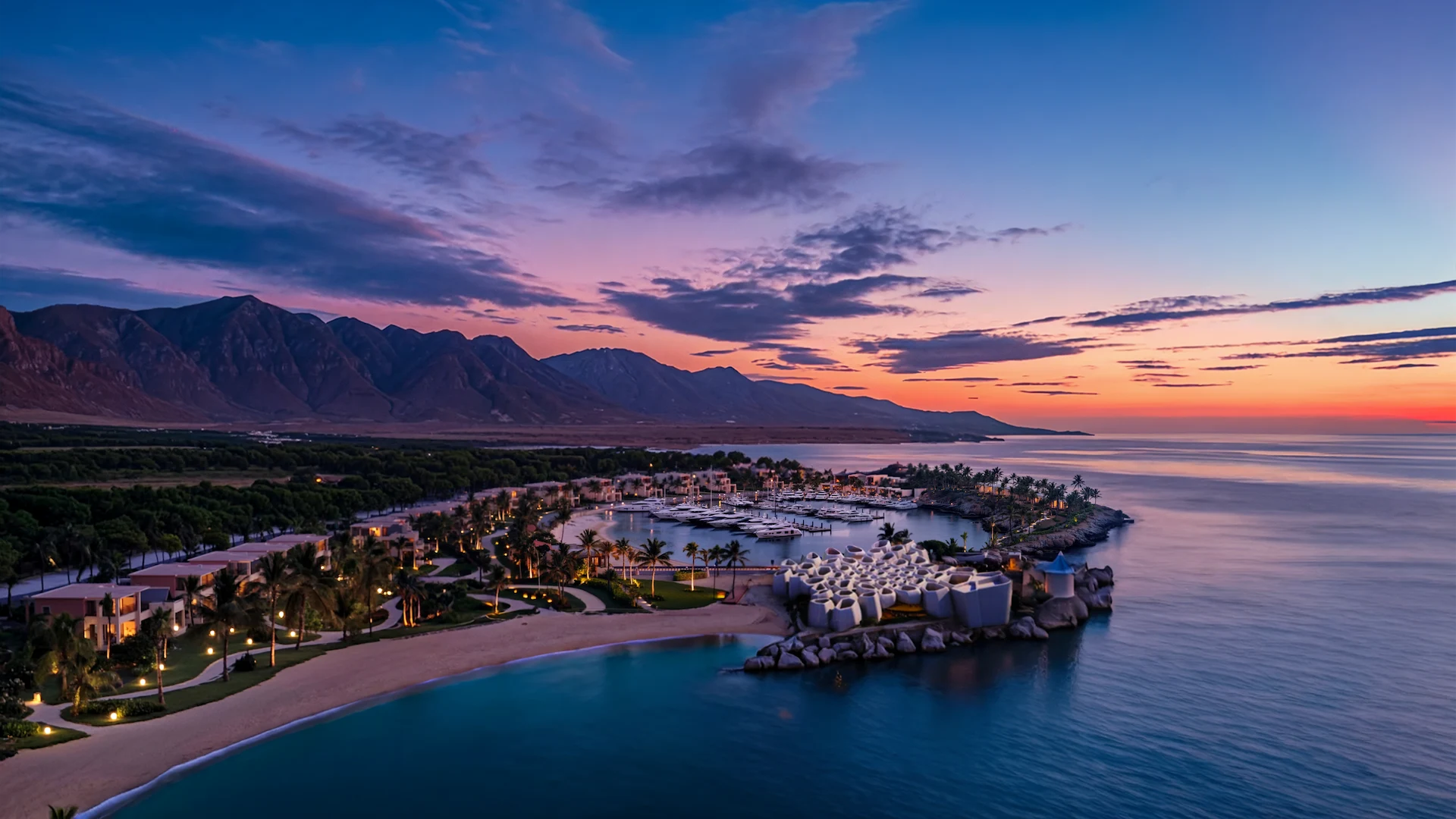 Sunset view of coastal resort with marina, yachts, palm-lined beach, and mountains in the background